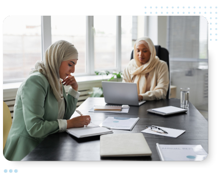 Two women in hijabs work at a desk with laptops and documents, focused and engaged. Office setting with large windows and a calm, professional atmosphere.
