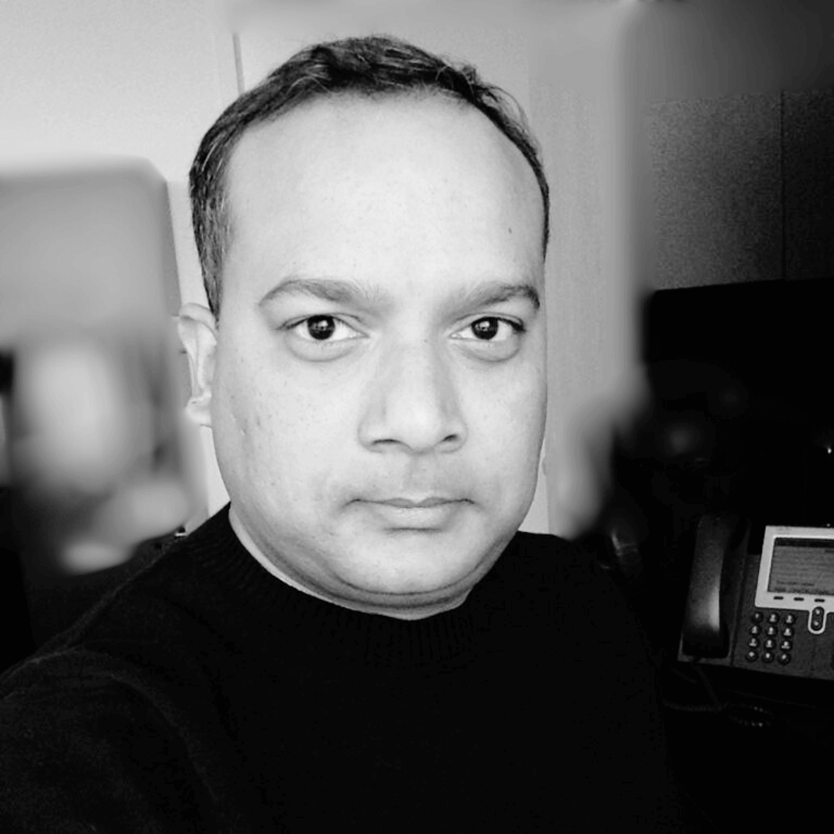 Black and white close-up of a person in a dark sweater, with short hair and a neutral expression, standing indoors near an office phone and blurred background.