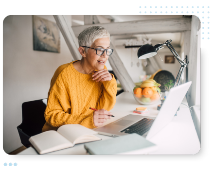 Senior woman with short gray hair in a cozy yellow sweater working on a laptop at a white desk, smiling thoughtfully with a notebook open nearby.